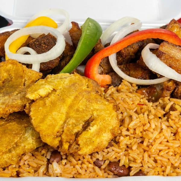 Takeout tray filled with seasoned rice and beans, fried plantains, grilled meat, onion rings, and red and green peppers.