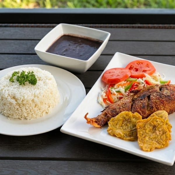 Fried whole fish with rice, vegetables, fried plantains, and dark dipping sauce served on a wooden outdoor table.