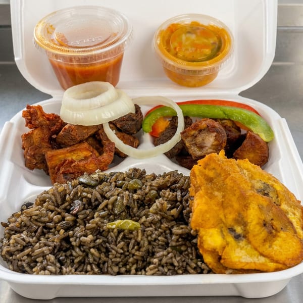 Takeout container holding seasoned rice, fried plantains, grilled meat, onions, peppers, and two cups of sauce.