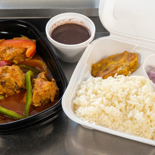 Takeout meal with stewed chicken and peppers, white rice, fried plantains, pickled onions, and a cup of dark sauce.